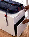 Person opening a white drawer mobile storage pedestal with a navy blue backpack on top. Kansas City Office Furniture.