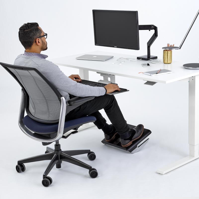 Person using a standing desk with a Humanscale FR500 black footrest in a modern office setting. Kansas City Office Design.