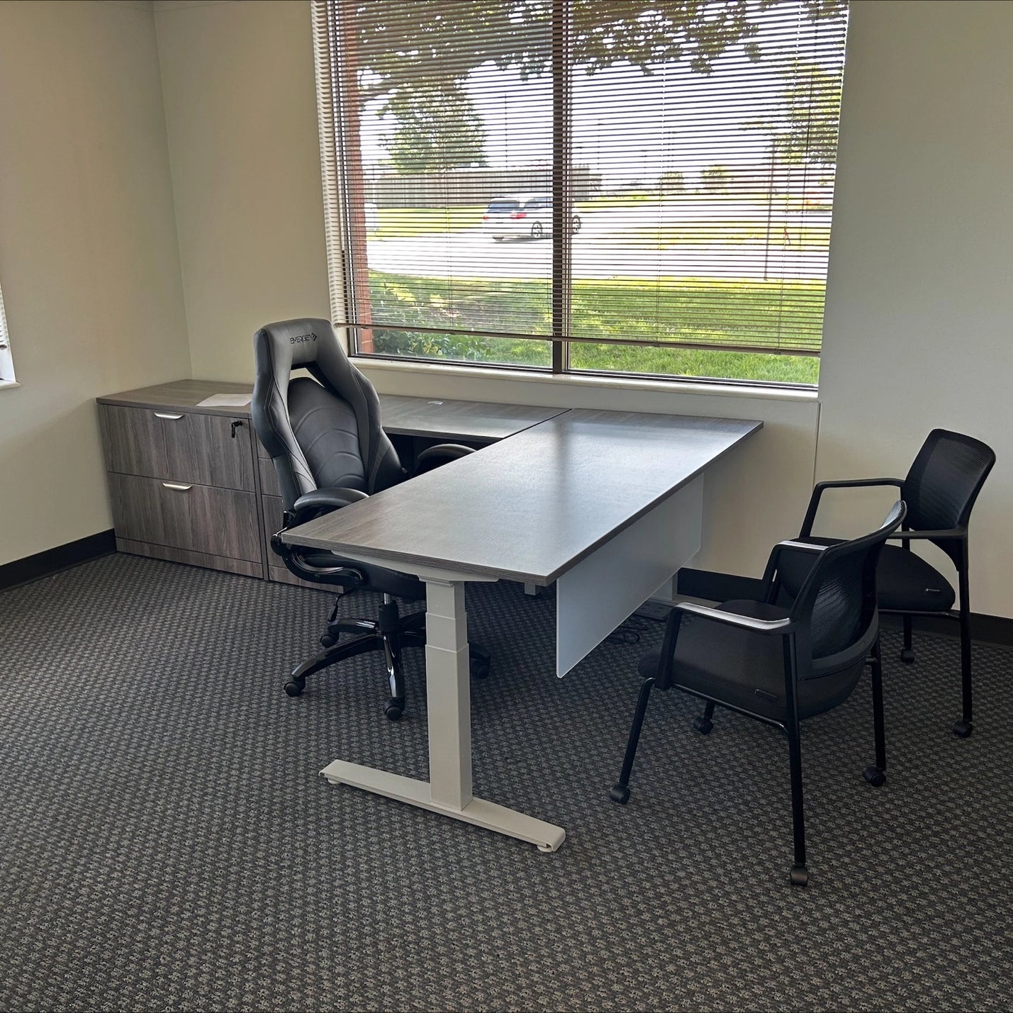 Office setup with a sit to stand height adjustable desk, chair, and additional chairs in front of a window.