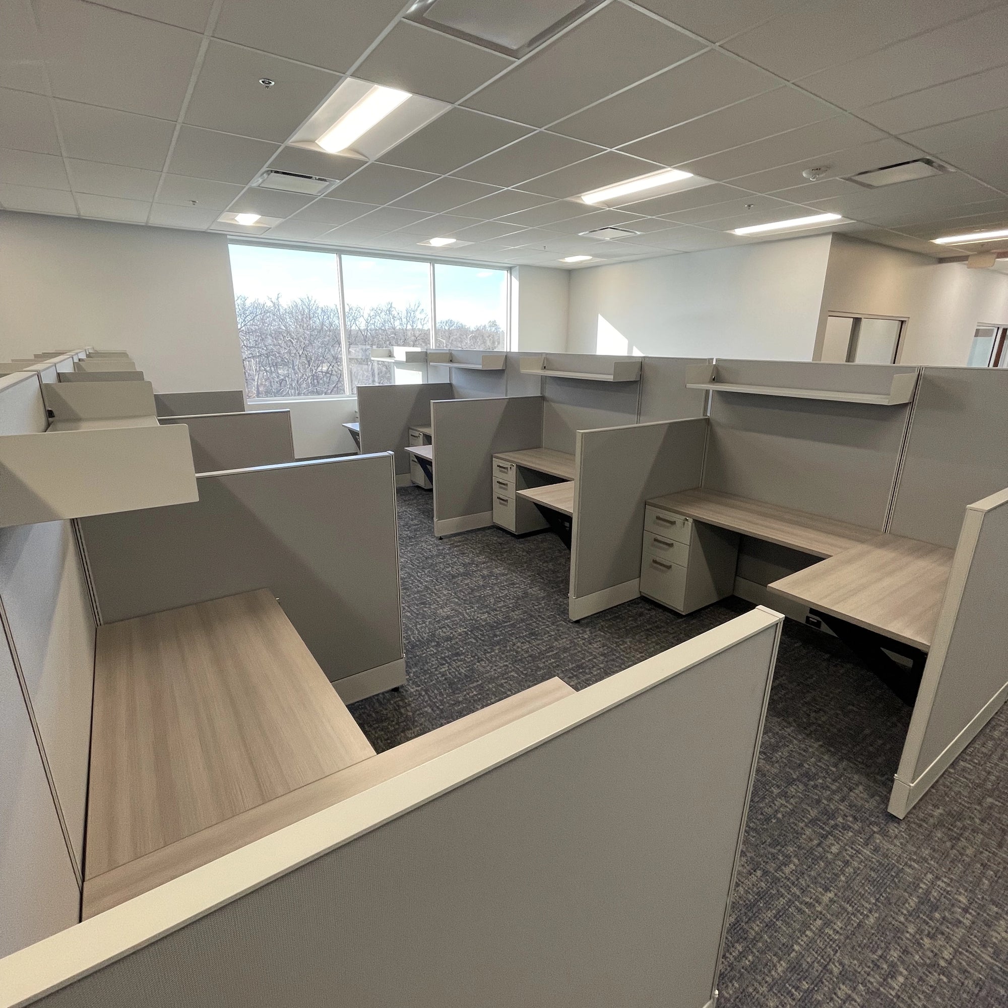 Modern office cubicles with large windows in a well-lit room. Orange Ev. Kansas City Office Design.