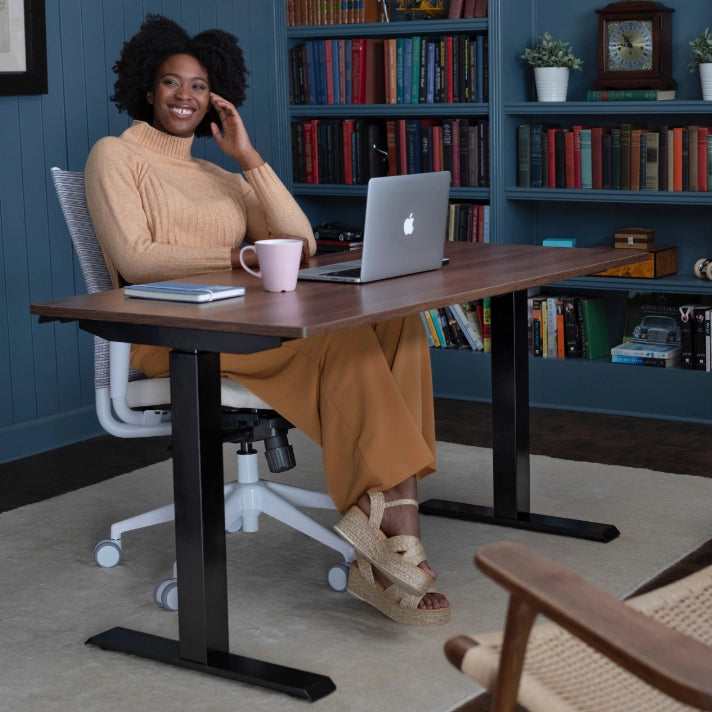 Woman sitting at a SitonIt Height Adjustable desk with a laptop and mug, surrounded by books in a home office setting. Kansas City Office Design.