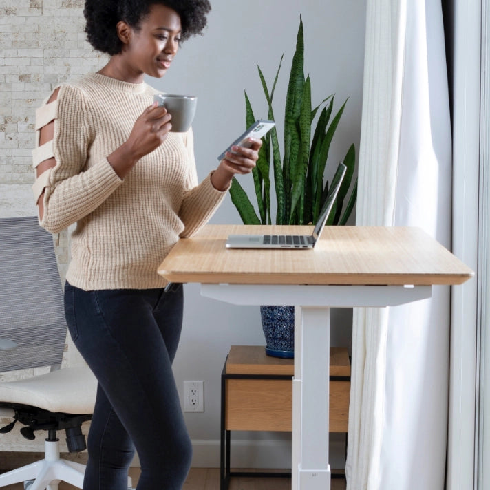 Woman using a smartphone next to a standing desk with a laptop and plant in a home office setting. Kansas City Office Design.