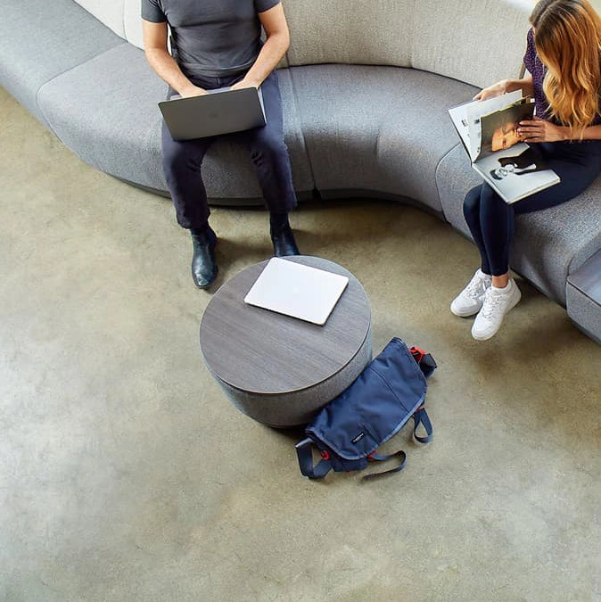 Two people sitting on a modern sofa with a round wooden table and a blue backpack on the floor. Kansas City Office Design.