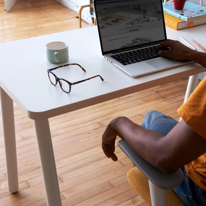Person sitting at a SitOnIt Reya desk with a laptop, mug, and glasses on a wooden floor. Kansas City Office Design.