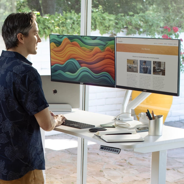 Person using a dual-monitor computer setup in a bright room with greenery outside. Kansas City Office Design.